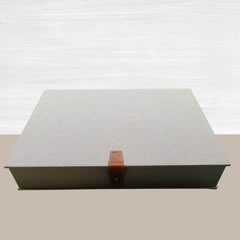 Un cofanetto vintage artigianale di forma rettangolare, rivestito in tessuto di canapa color beige naturale. La scatola è chiusa da un raffinato cinturino in vera pelle marrone chiaro con un bottone a pressione metallico argentato.