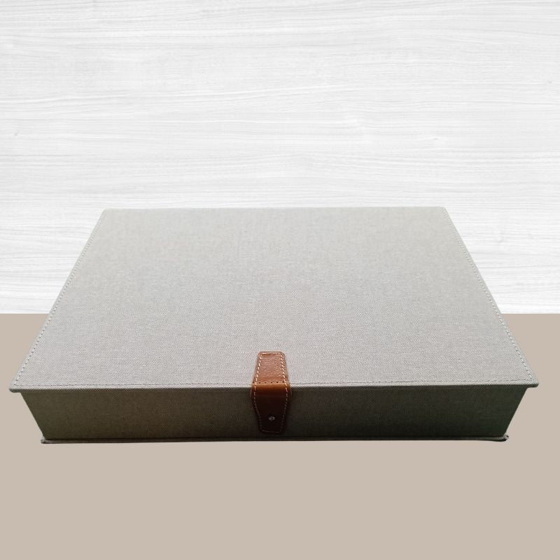 Un cofanetto vintage artigianale di forma rettangolare, rivestito in tessuto di canapa color beige naturale. La scatola è chiusa da un raffinato cinturino in vera pelle marrone chiaro con un bottone a pressione metallico argentato.